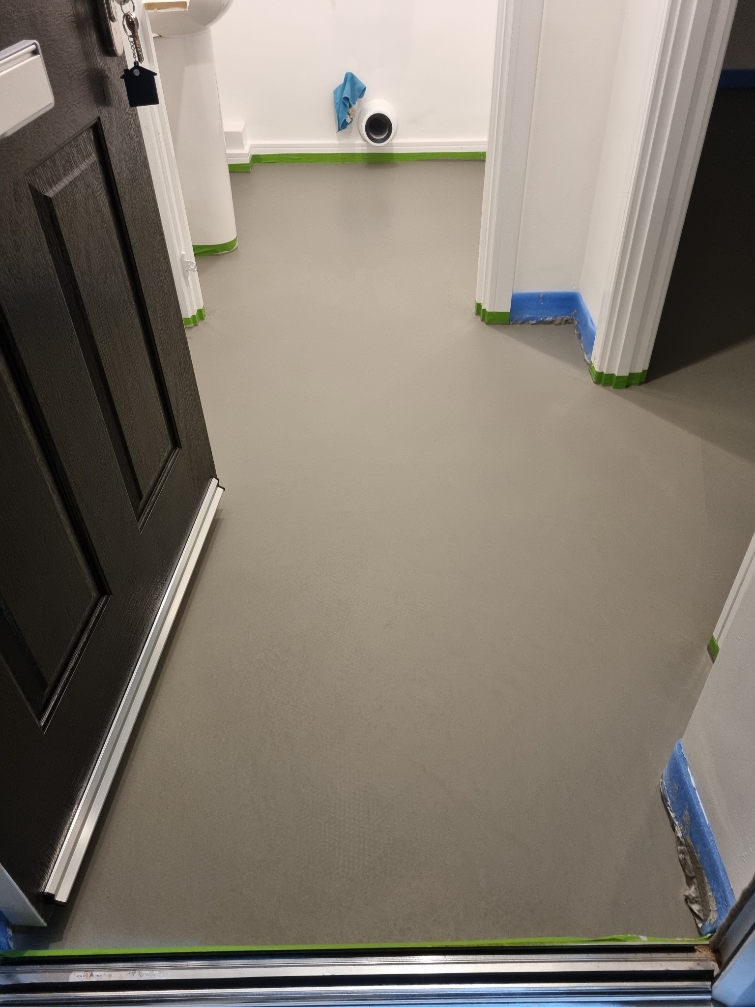 Świeżo wylana, szara posadzka żywiczna w trakcie schnięcia, zabezpieczona taśmą malarską przy ścianach i ościeżnicach drzwiowych, widok zza uchylonych, ciemnych drzwi wejściowych.