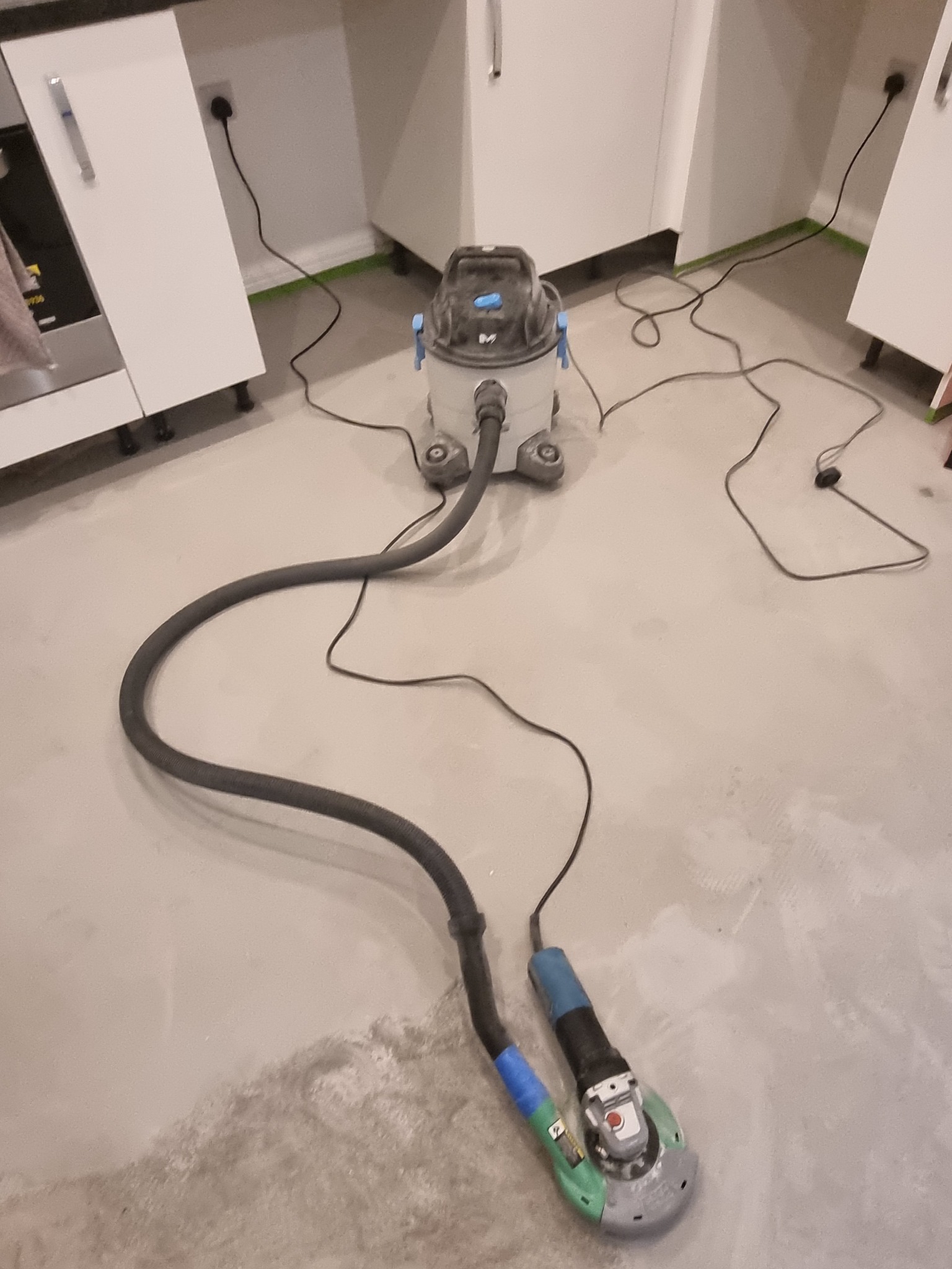 Szlifowanie podłogi w kuchni podczas remontu, szlifierka kątowa podłączona do odkurzacza przemysłowego, widoczne ślady po szlifowaniu na podłodze, zabezpieczone dolne krawędzie szafek kuchennych.