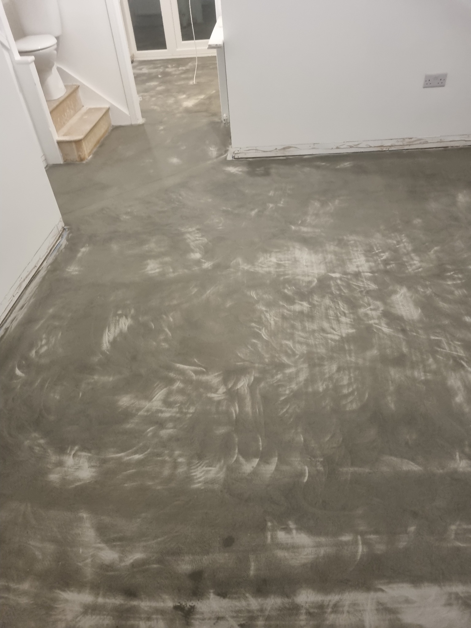 Świeżo wylana, szara, samopoziomująca wylewka podłogowa w trakcie schnięcia, widoczne ślady po rozprowadzaniu masy, białe ściany w tle, przygotowane do dalszych prac wykończeniowych.