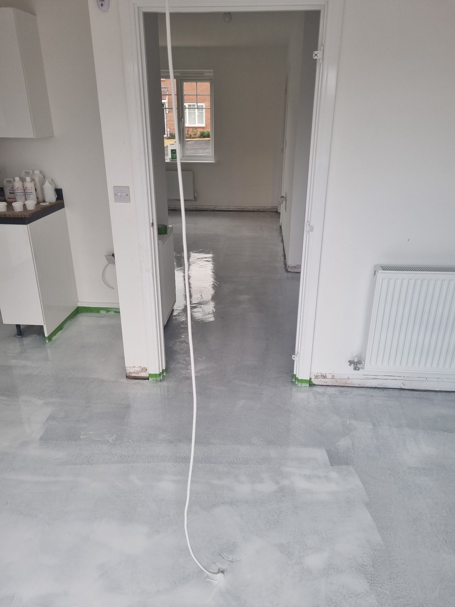 Świeżo wylana, błyszcząca posadzka w jasnym pomieszczeniu z widocznymi śladami po wałku, zabezpieczona taśmą malarską przy ścianach i drzwiach, z widocznymi butelkami z preparatami na blacie...