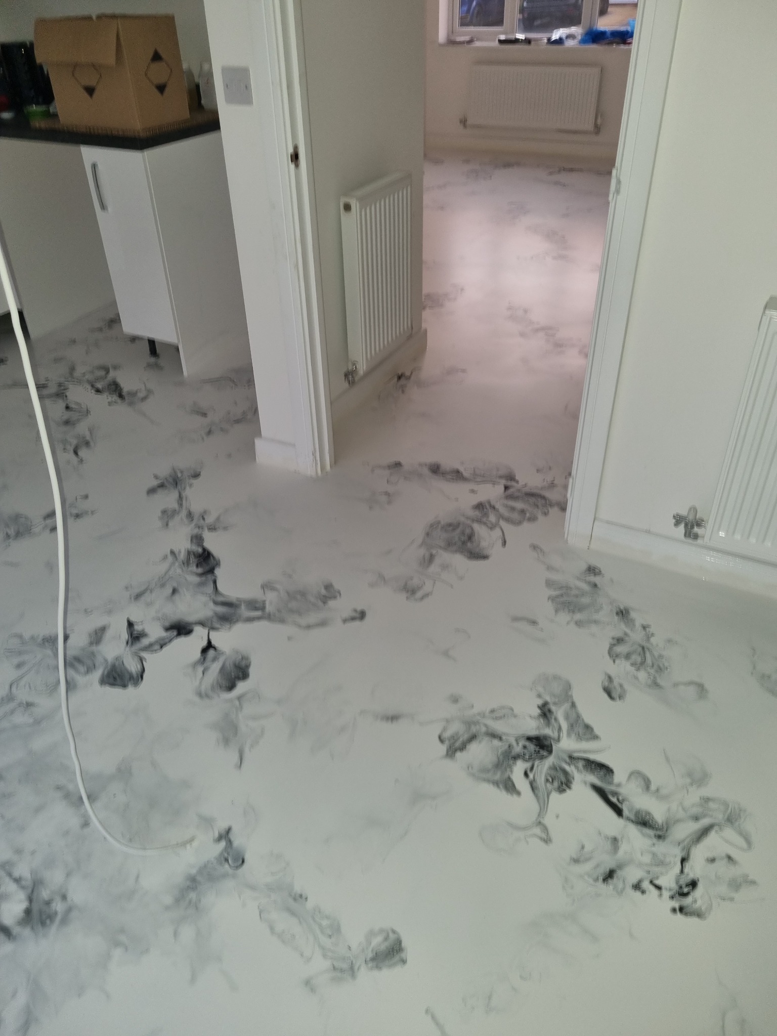 Biała podłoga żywiczna z szarymi wzorami przypominającymi marmur, widok z perspektywy, fragment szafki kuchennej i grzejniki w tle.