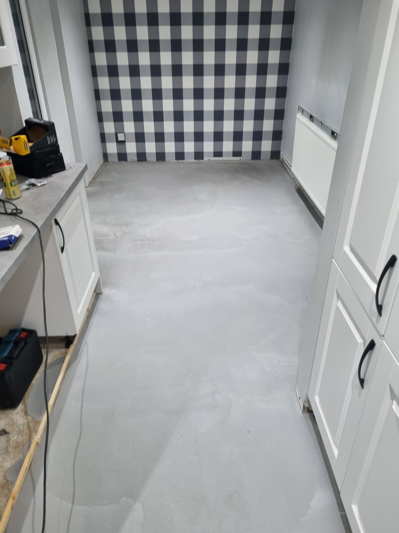 Wnętrze kuchni w trakcie prac wykończeniowych; widać świeżo wylany, jasnoszary betonowy podkład podłogowy, białe szafki kuchenne z czarnymi uchwytami, narzędzia na blacie i tapetę w czarno-białą...