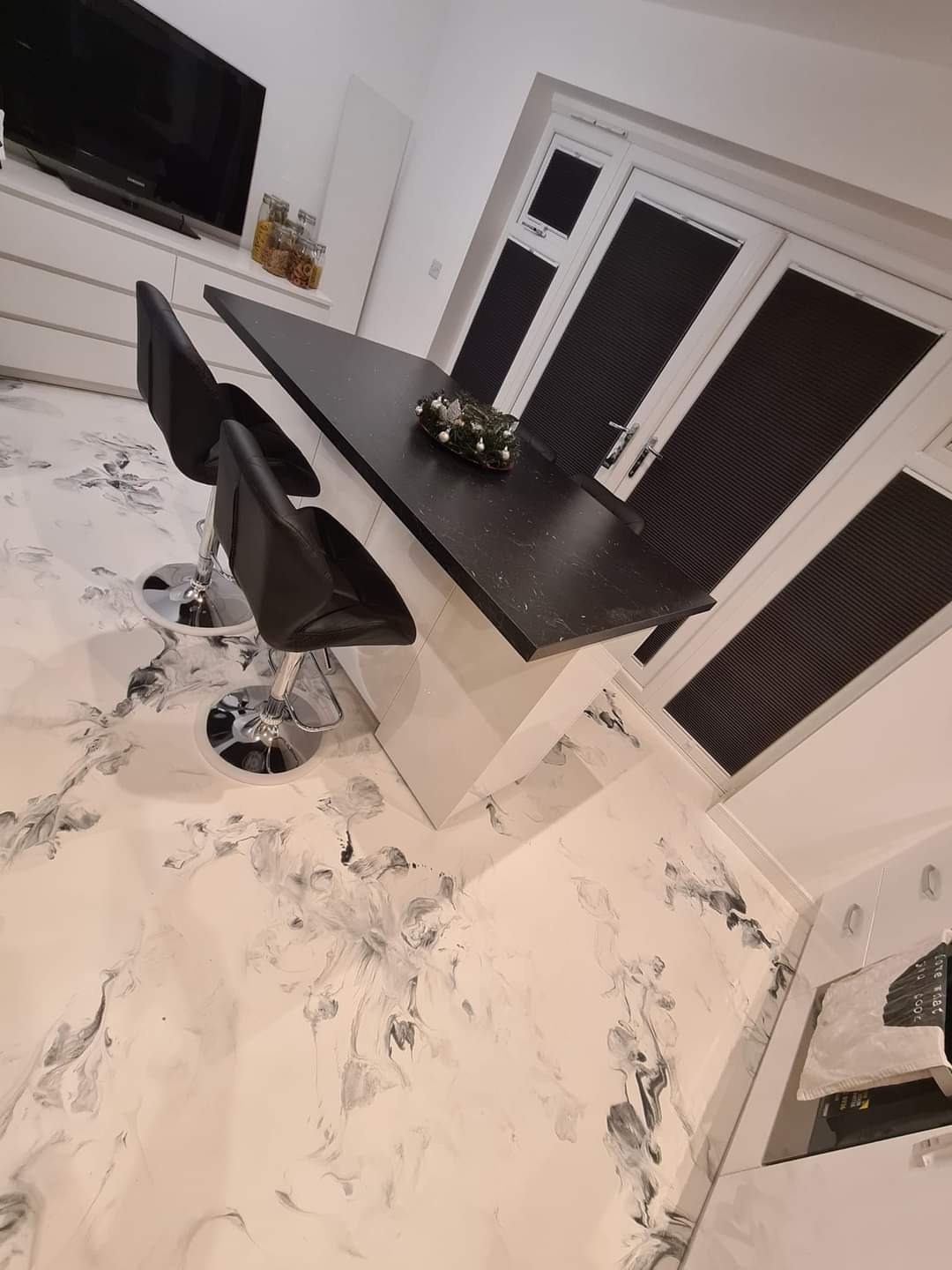 Nowoczesna kuchnia z wyspą i czarnymi hokerami, podłoga w stylu marmuru, okna z żaluzjami plisowanymi, widok z perspektywy podłogi.