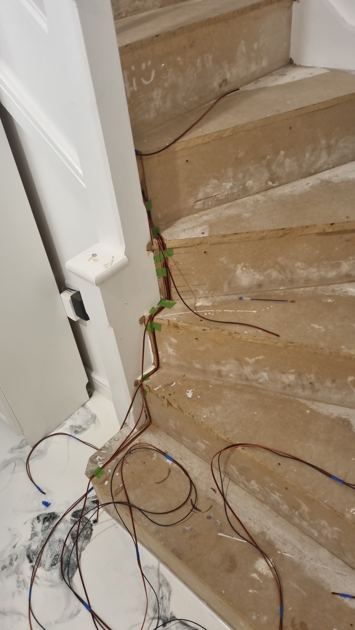 Surowe drewniane schody w trakcie instalacji oświetlenia, z widocznymi przewodami przytwierdzonymi taśmą malarską do ściany i schodów. Na ścianie obok schodów widoczny biały uśmiech narysowany...