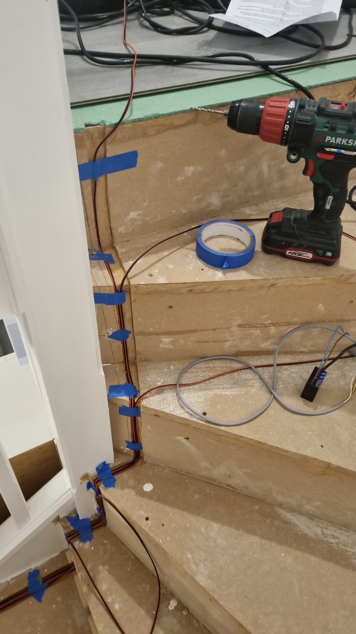 Montaż oświetlenia LED na schodach w trakcie prac: przewody przymocowane niebieską taśmą malarską, wiertarka akumulatorowa Parkside i rolka taśmy leżą na drewnianych stopniach.