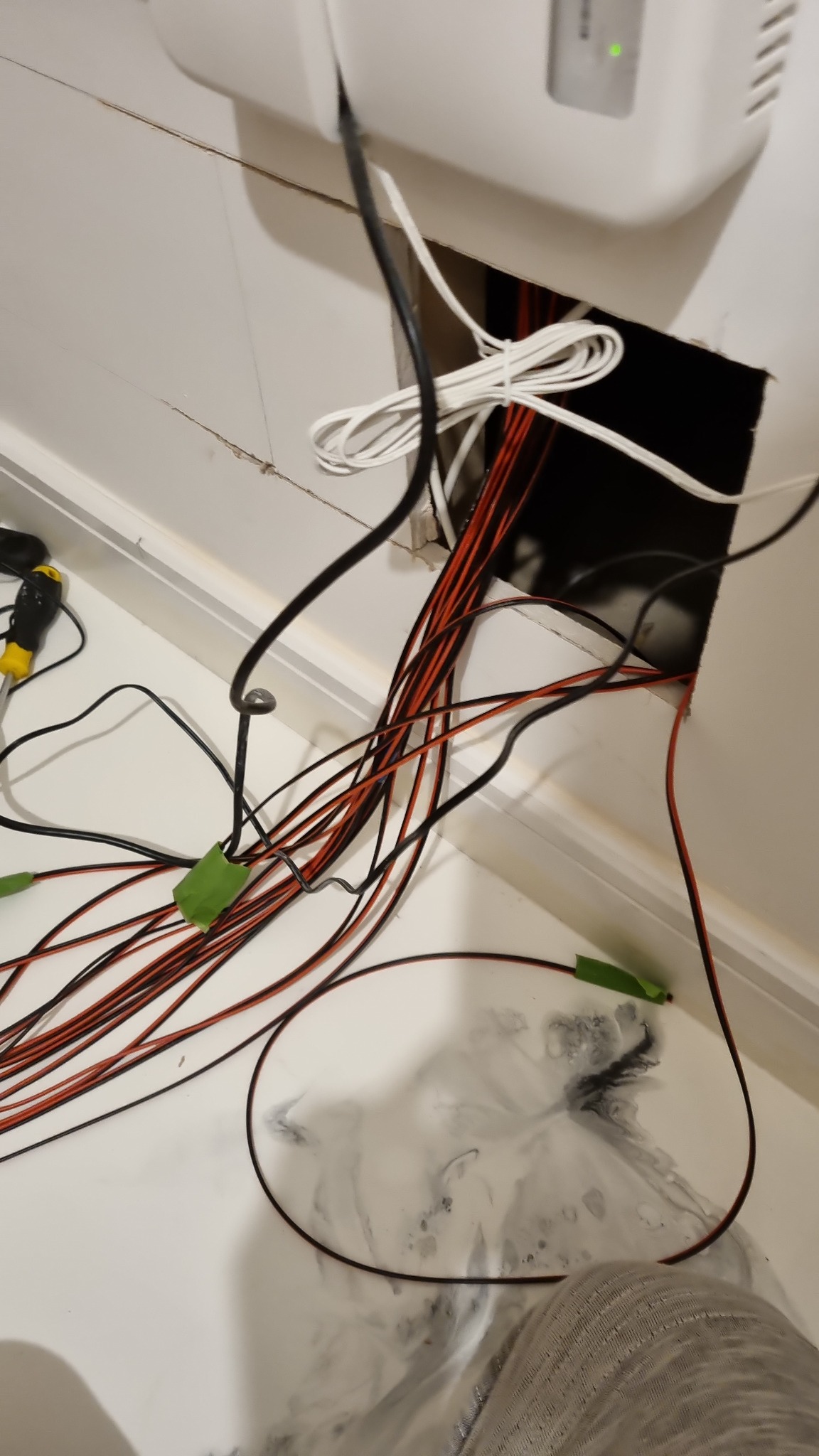 Niedokończone podłączenie przewodów elektrycznych wychodzących z otworu w suficie, widoczne przewody czarne i czerwone, narzędzia i ślady po wierceniu na podłodze.