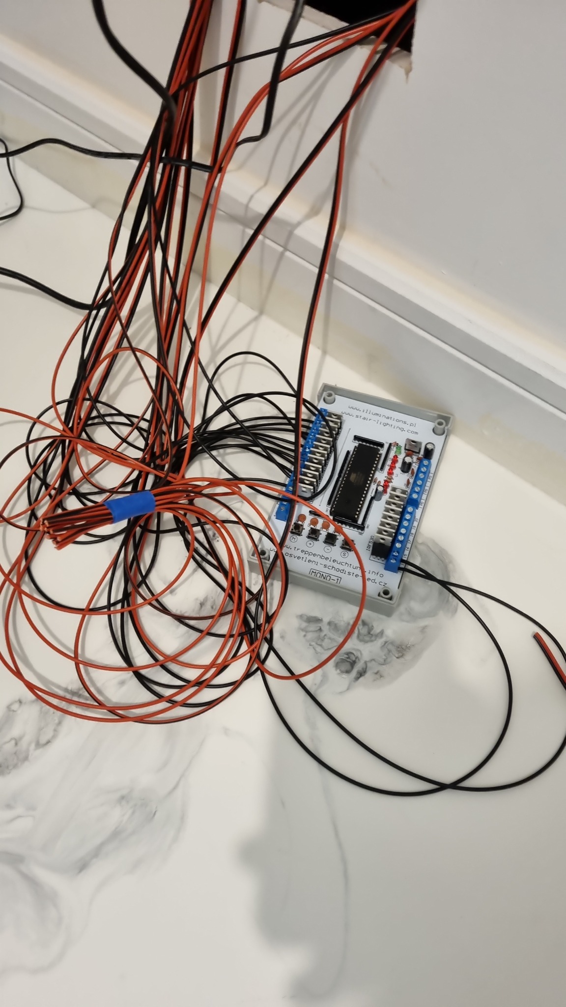 Rozplątane czarne i czerwone przewody elektryczne, część związana niebieską taśmą, prowadzące do płytki sterującej oświetleniem schodowym MONO-1, leżącej na marmurowym blacie.