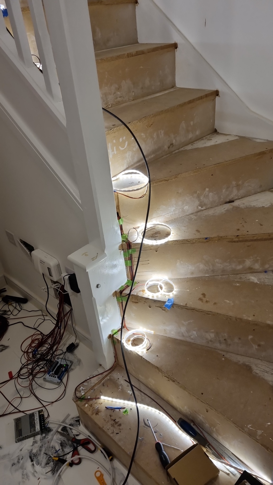 Montaż oświetlenia LED na schodach w trakcie remontu, widoczne przewody, taśmy montażowe, narzędzia i nieukończone stopnie.
