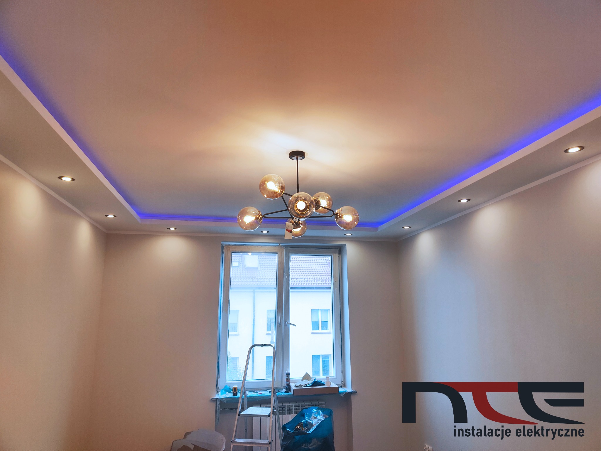 Nowoczesne oświetlenie sufitowe z listwami LED RGB i lampą wiszącą z kulistymi kloszami, widok na okno z drabiną i workiem na śmieci, logo firmy na ścianie.