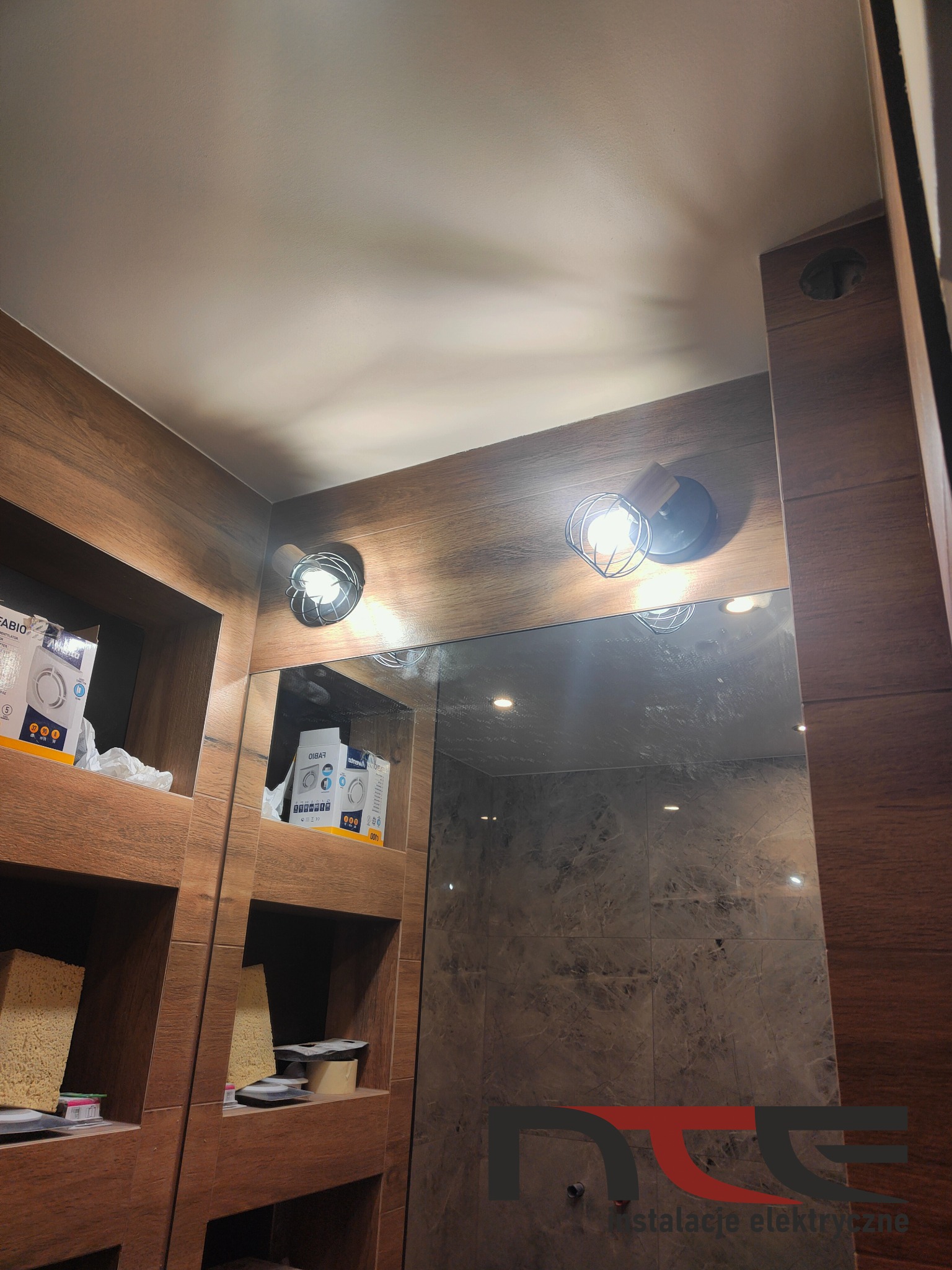 Łazienka z drewnianymi elementami, oświetleniem punktowym typu Edison oraz lustrem odbijającym marmurowe płytki w kabinie prysznicowej. Na półkach widoczne opakowania wentylatorów.