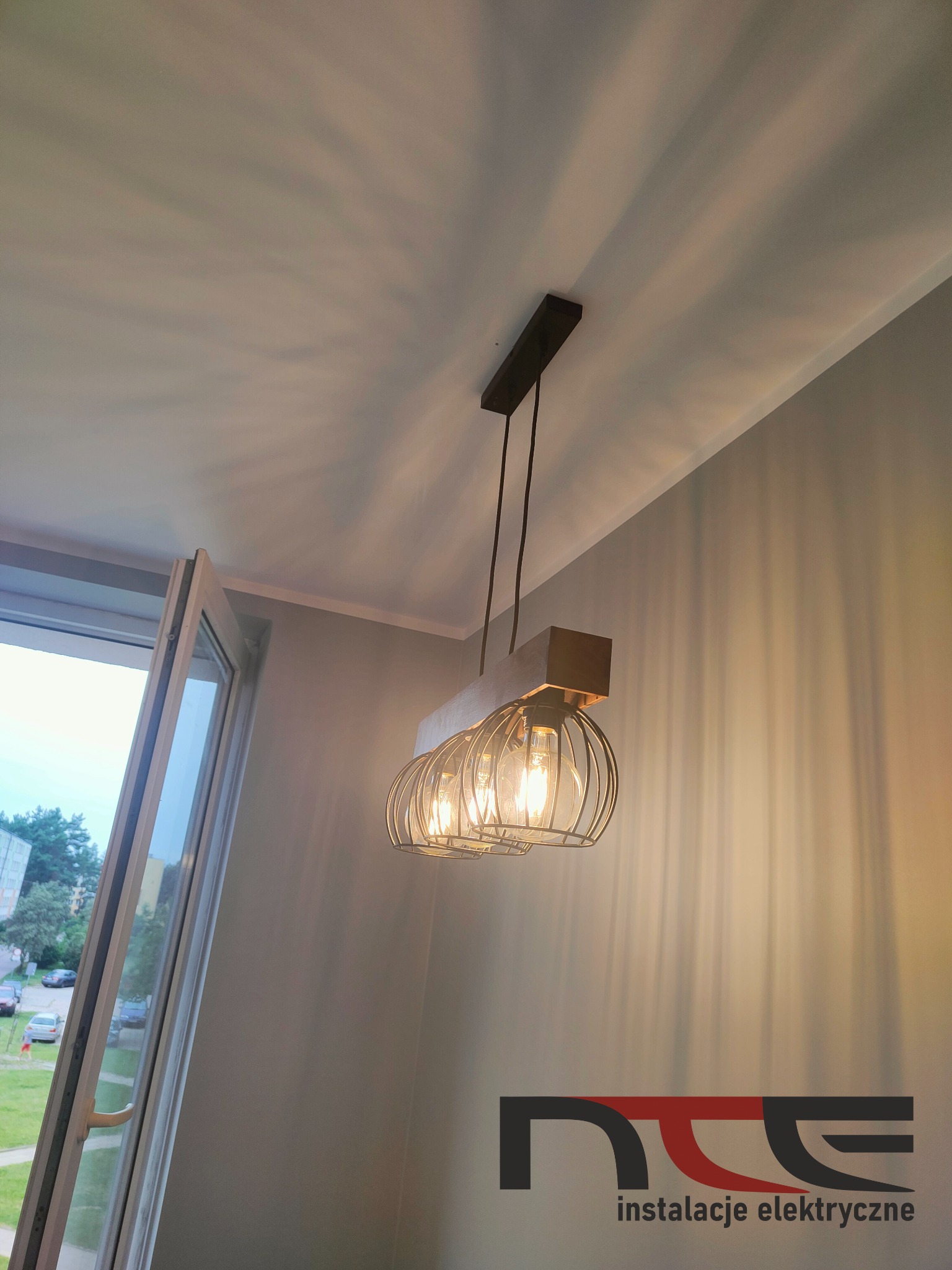 Zawieszona, metalowa lampa klatkowa z trzema żarówkami Edisona, przymocowana do drewnianej belki, oświetlająca szarą ścianę i sufit w pokoju z otwartym oknem.