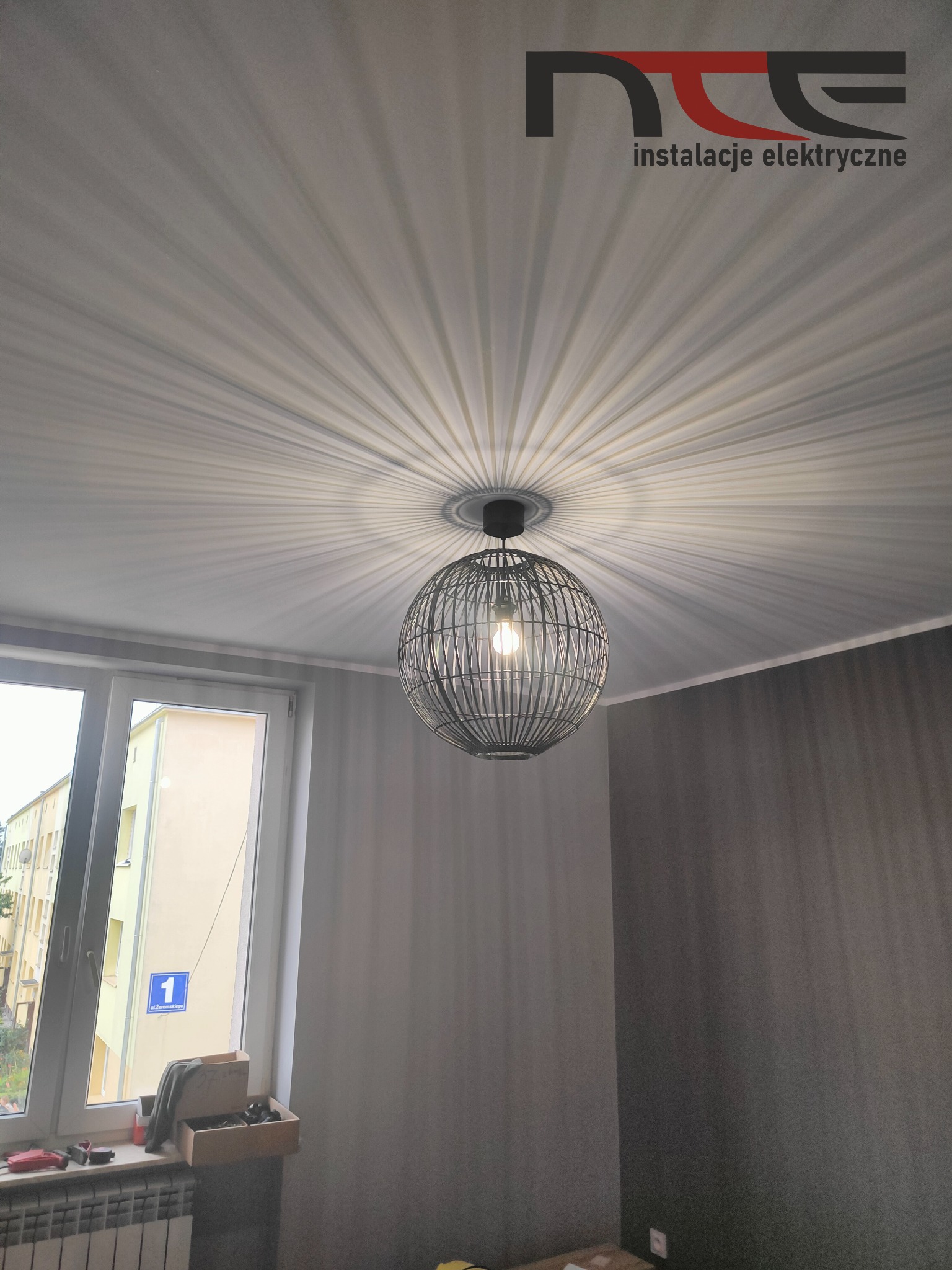 Czarna, druciana lampa wisząca w kształcie kuli rzuca promieniujące cienie na biały sufit w pokoju z szarą ścianą. W tle okno z widokiem na budynek z adresem 'ul. Żeromskiego 1'. W rogu widoczne...