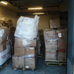 Magazyn z paletami kartonowych pudeł i dużym białym workiem, niektóre pudła opisane 'Nappies', widoczne przez otwarte drzwi magazynu.