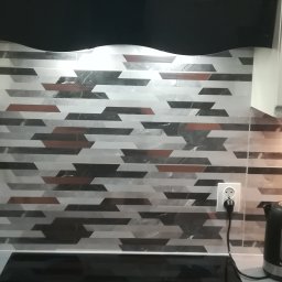 Marcin Rozlepiło - Wzorzyste płytki ścienne w kuchni, ułożone poziomo w pasy z motywem geometrycznym w odcieniach szarości, czerni i brązu, widoczny fragment płyty indukcyjnej i czajnika elektrycznego oraz gniazdko...