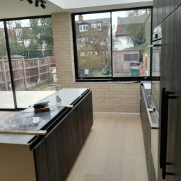 Marcin Rozlepiło - Wnętrze w trakcie montażu kuchni z ciemnymi, drewnopodobnymi frontami i jasną podłogą, widok na ogród przez duże okna z czarnymi ramami, sufit z oświetleniem punktowym.