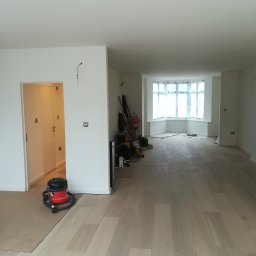 Marcin Rozlepiło - Jasne panele podłogowe ułożone w przestronnym, odnowionym wnętrzu z oknem wykuszowym zabezpieczonym folią malarską, widoczny odkurzacz i narzędzia malarskie.