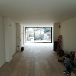 Marcin Rozlepiło - Jasne, przestronne wnętrze po remoncie z drewnianą podłogą, świeżo pomalowanymi ścianami i dużym oknem panoramicznym wychodzącym na ogród. Na podłodze widoczne narzędzia i akcesoria malarskie...