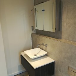 Marcin Rozlepiło - Nowoczesna łazienka w trakcie wykańczania: biała umywalka nablatowa, szafka wisząca z ciemnego drewna, lustro z oświetleniem LED, szare płytki na ścianie, widoczne kable elektryczne i narzędzia.