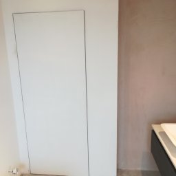 Marcin Rozlepiło - Ukryte drzwi rewizyjne w łazience, ściana po szpachlowaniu przed malowaniem, widoczna zabudowa umywalki i fragment płytek podłogowych.