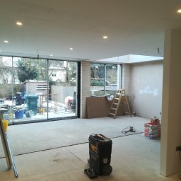 Marcin Rozlepiło - Wnętrze w trakcie remontu: szpachlowane ściany, widoczna drabina, worki z zaprawą, skrzynka narzędziowa Stanley i duże okno z widokiem na zasypany śniegiem ogród.