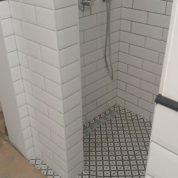 Marcin Rozlepiło - Wykończona kabina prysznicowa z białymi, prostokątnymi płytkami ułożonymi w cegiełkę i wzorzystą, czarno-białą podłogą z odpływem liniowym.