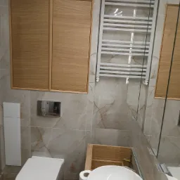 Nowoczesna łazienka z beżowymi płytkami imitującymi marmur, wisząca toaleta, drewniane szafki, biały grzejnik drabinkowy i umywalka z baterią.