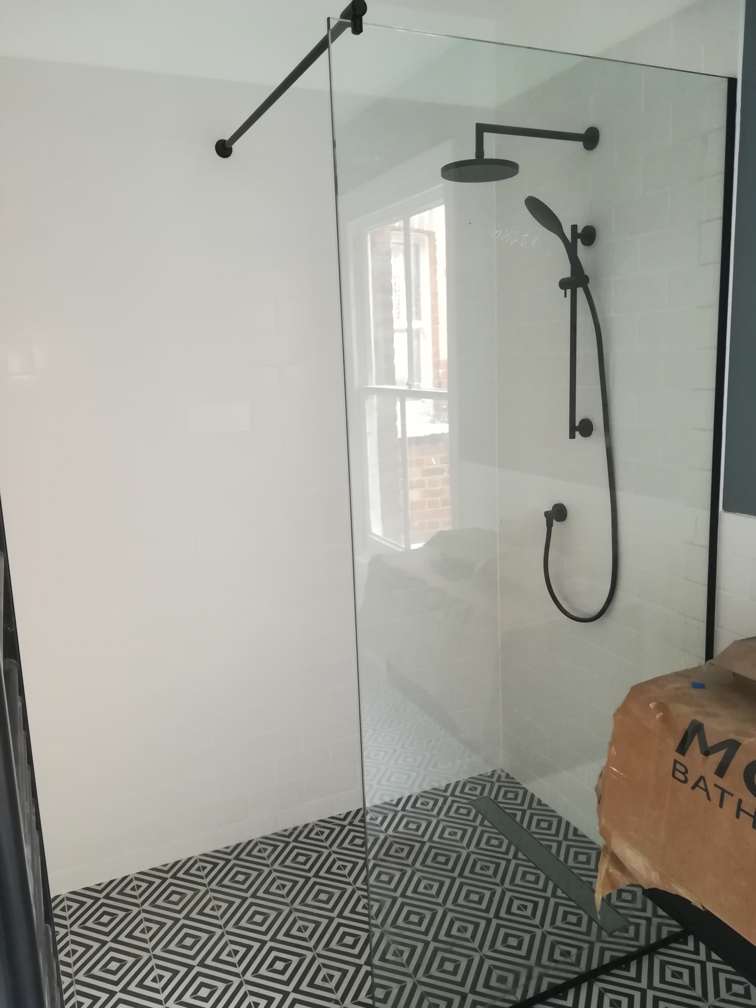 Nowoczesna kabina prysznicowa z czarną armaturą i geometryczną posadzką, widok z boku, częściowo zakryta kartonem z napisem 'BATH'.