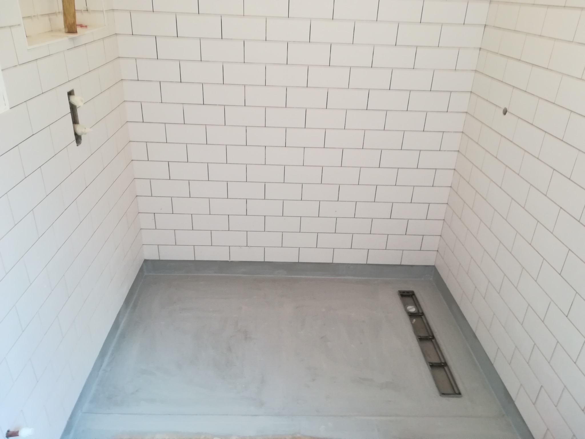 Przygotowany brodzik z odpływem liniowym i częściowo wyłożonymi białymi, prostokątnymi płytkami ścianami. Widoczne otwory na armaturę.