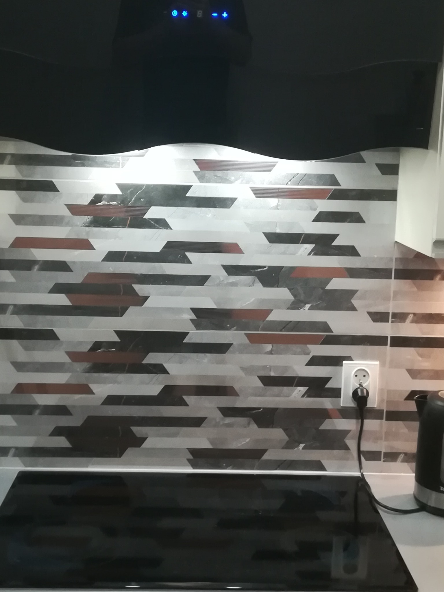 Wzorzyste płytki ścienne w kuchni, ułożone poziomo w pasy z motywem geometrycznym w odcieniach szarości, czerni i brązu, widoczny fragment płyty indukcyjnej i czajnika elektrycznego oraz gniazdko...