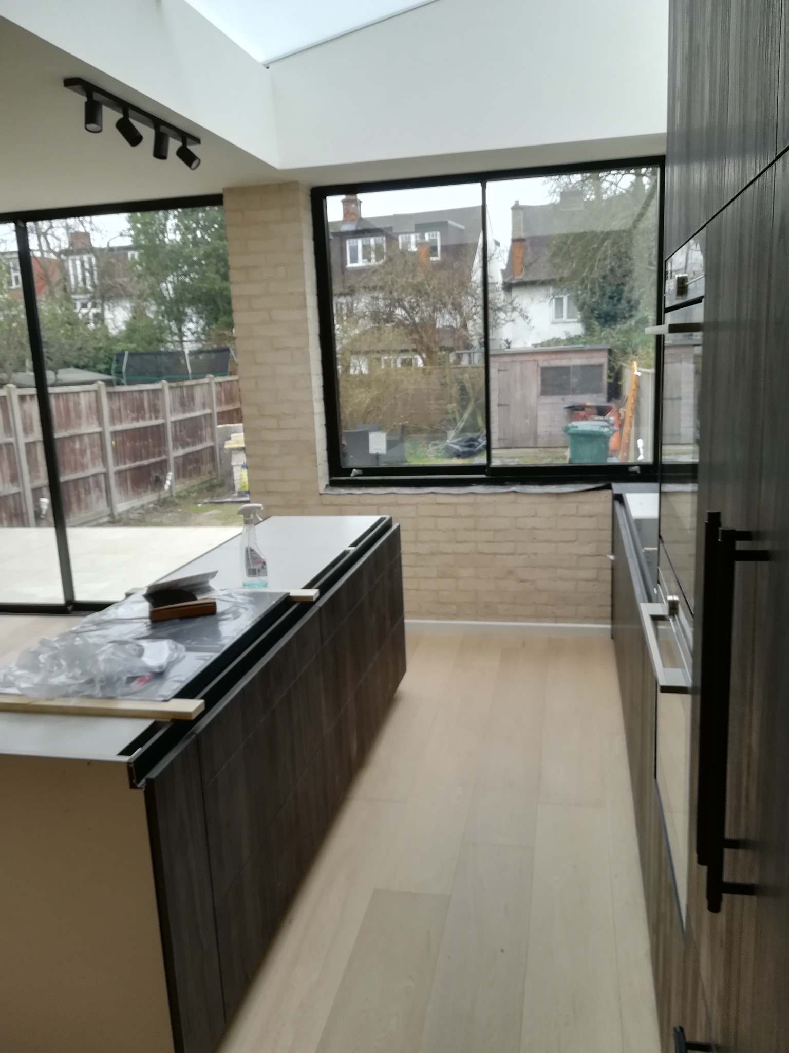 Wnętrze w trakcie montażu kuchni z ciemnymi, drewnopodobnymi frontami i jasną podłogą, widok na ogród przez duże okna z czarnymi ramami, sufit z oświetleniem punktowym.