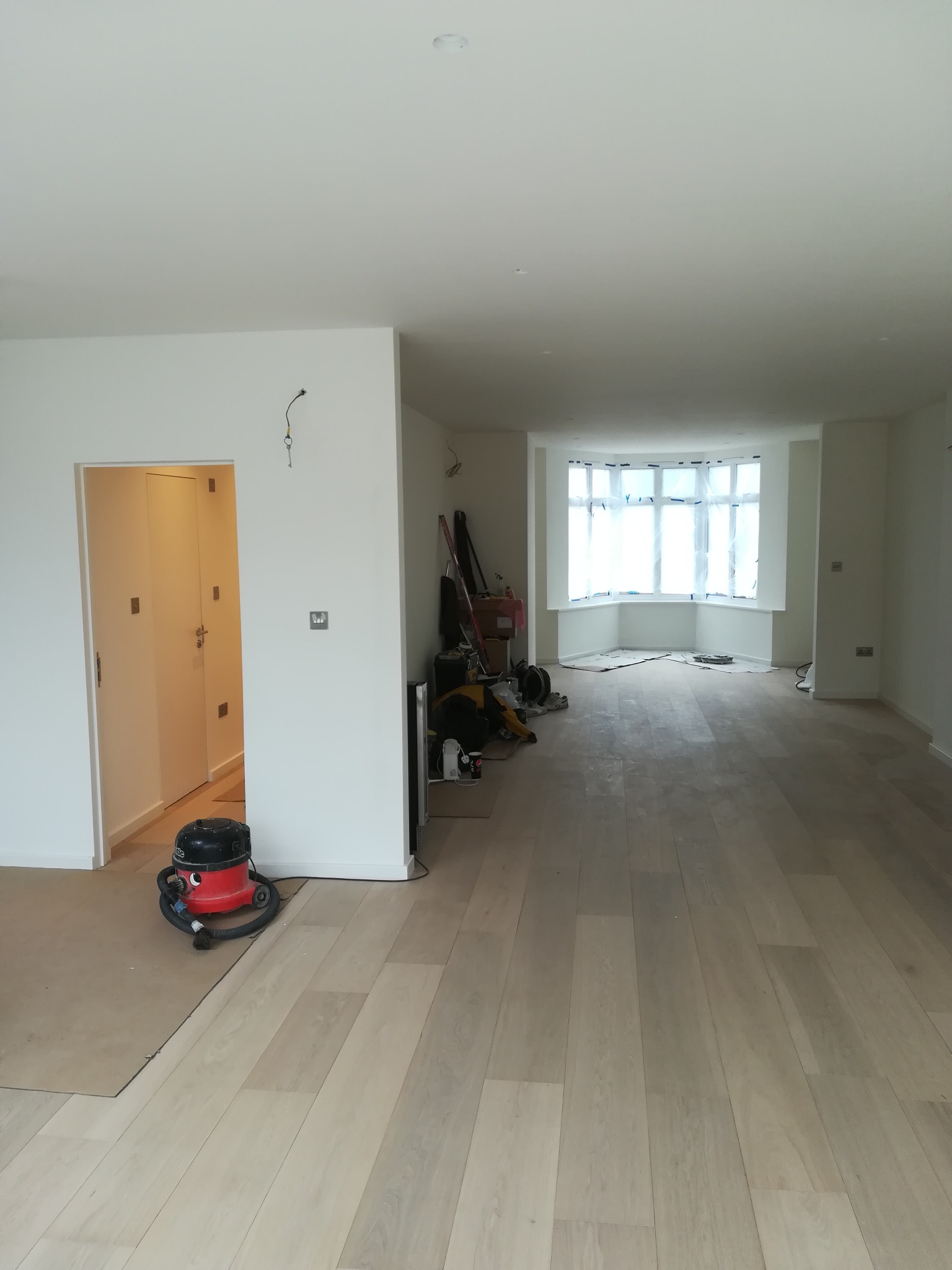 Jasne panele podłogowe ułożone w przestronnym, odnowionym wnętrzu z oknem wykuszowym zabezpieczonym folią malarską, widoczny odkurzacz i narzędzia malarskie.