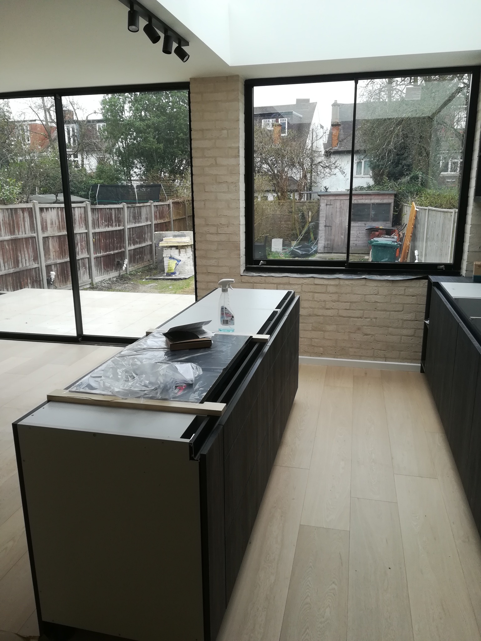 Wnętrze w trakcie prac wykończeniowych: widok na wyspę kuchenną z zabezpieczonym blatem, jasne panele podłogowe, ceglana ściana i duże okna z widokiem na ogród.