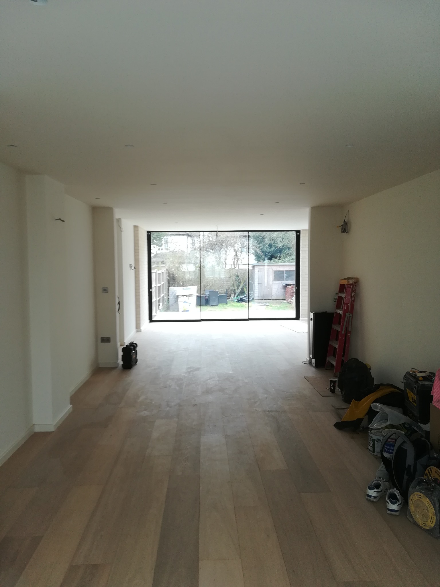 Jasne, przestronne wnętrze po remoncie z drewnianą podłogą, świeżo pomalowanymi ścianami i dużym oknem panoramicznym wychodzącym na ogród. Na podłodze widoczne narzędzia i akcesoria malarskie...