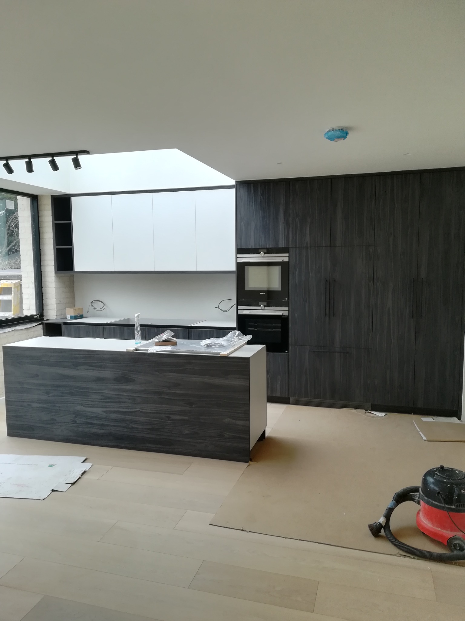 Nowoczesna kuchnia w trakcie montażu: wyspa z szarym fornirem, zabudowa kuchenna z ciemnego drewna z wbudowanym piekarnikiem i mikrofalówką Siemens, białe szafki wiszące pod świetlikiem, podłoga...