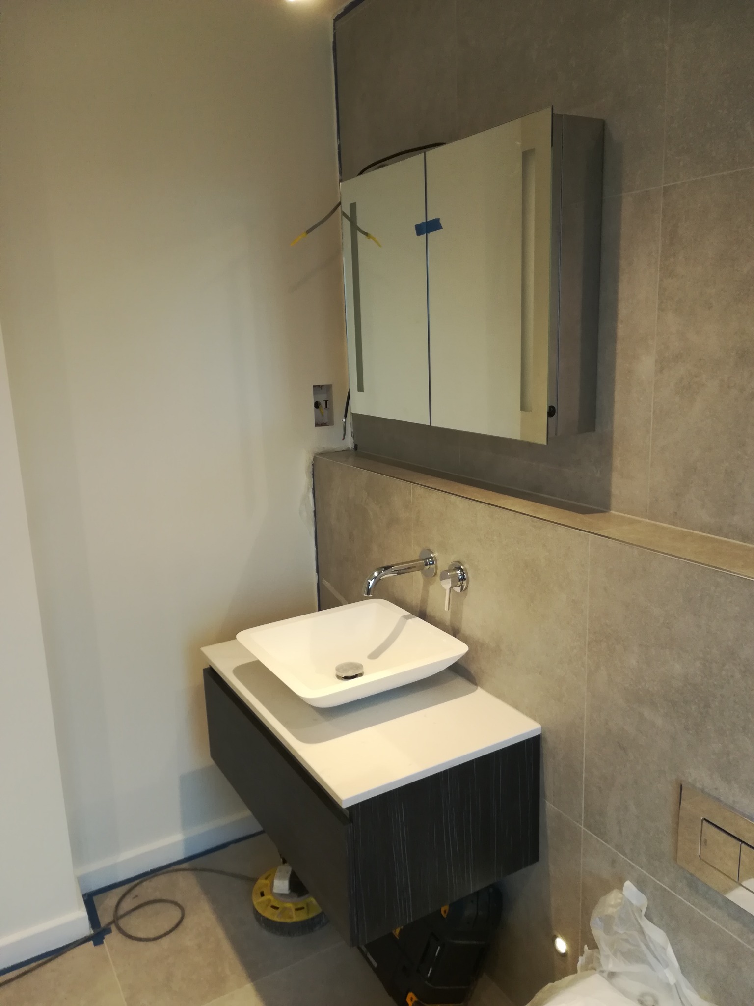 Nowoczesna łazienka w trakcie wykańczania: biała umywalka nablatowa, szafka wisząca z ciemnego drewna, lustro z oświetleniem LED, szare płytki na ścianie, widoczne kable elektryczne i narzędzia.