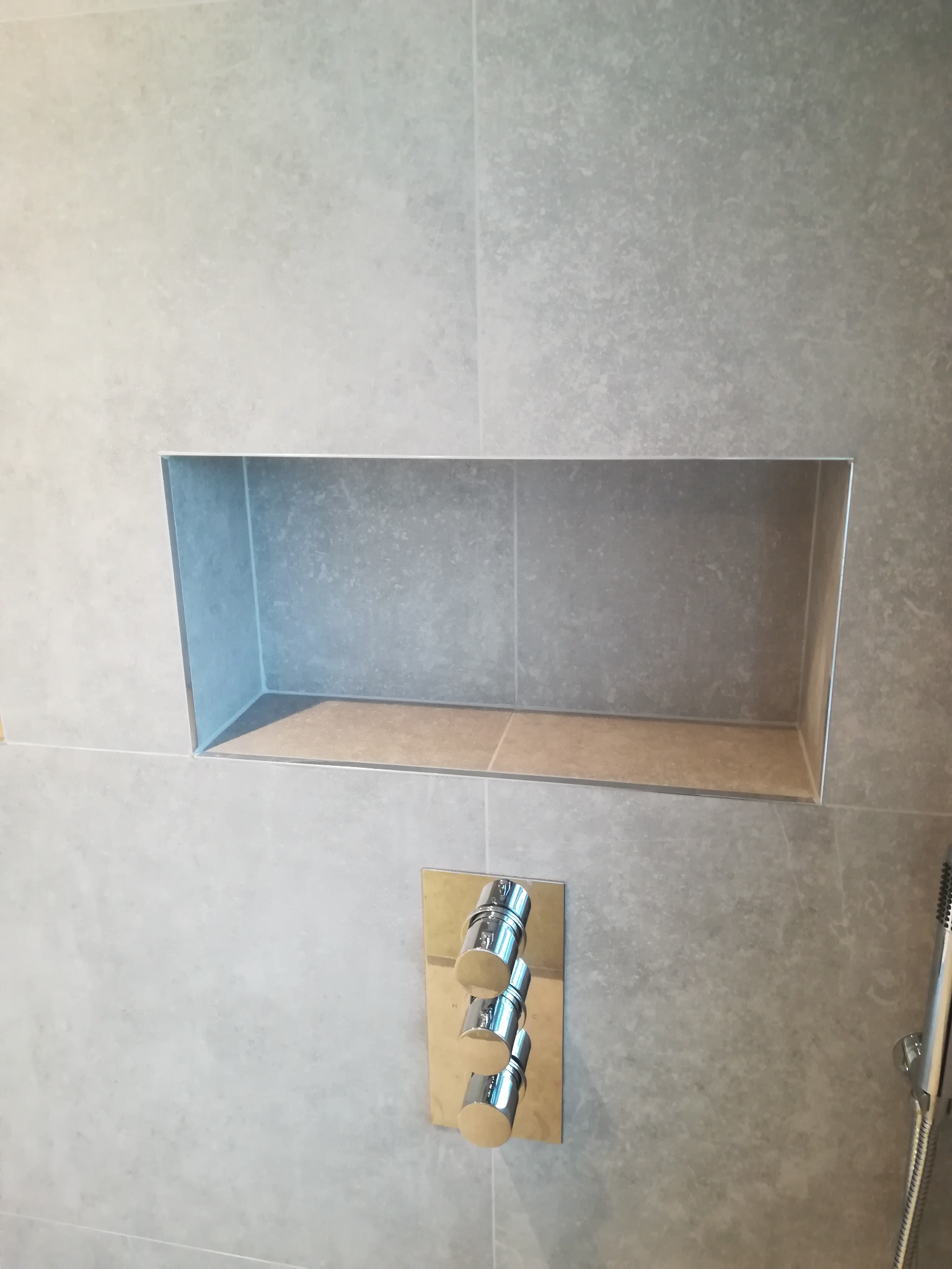 Szara ściana w łazience z wnęką wyłożoną płytkami i chromowaną baterią prysznicową z trzema pokrętłami.