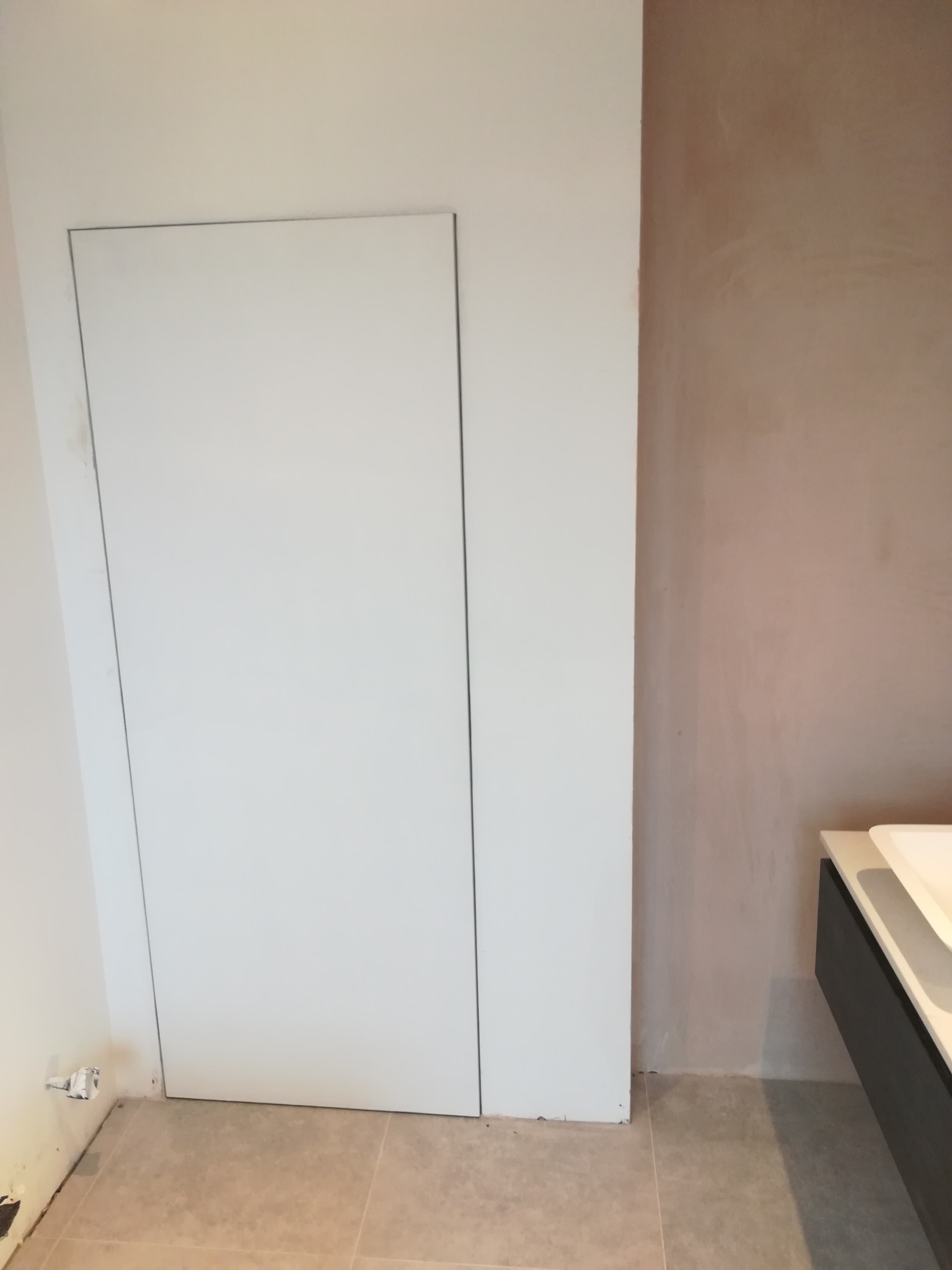 Ukryte drzwi rewizyjne w łazience, ściana po szpachlowaniu przed malowaniem, widoczna zabudowa umywalki i fragment płytek podłogowych.