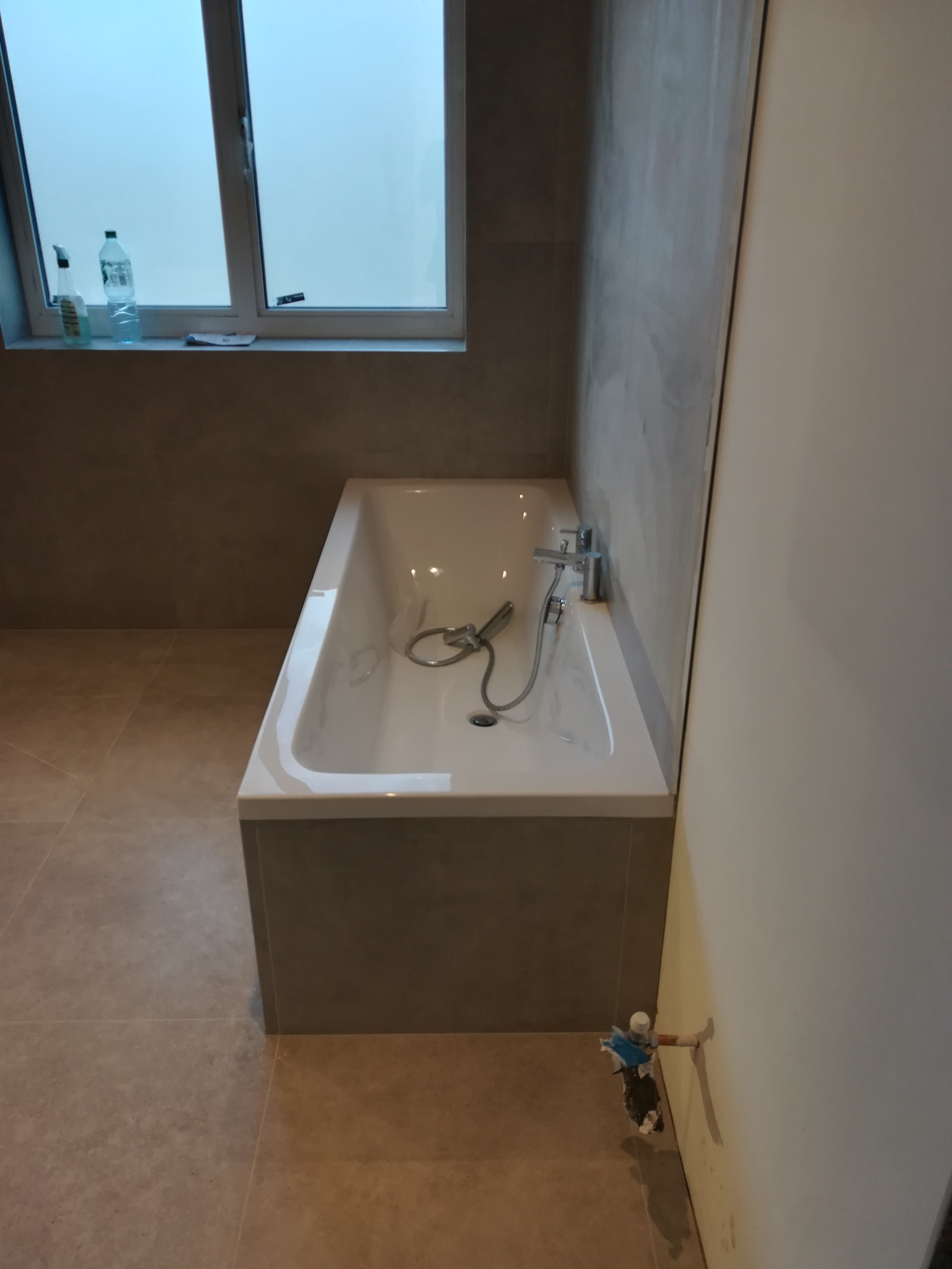 Nowoczesna wanna w obudowie z szarej płyty, zamontowana bateria z prysznicem, okno z mleczną szybą nad wanną, widoczne przyłącze wodne do baterii, szare płytki na ścianach i podłodze.