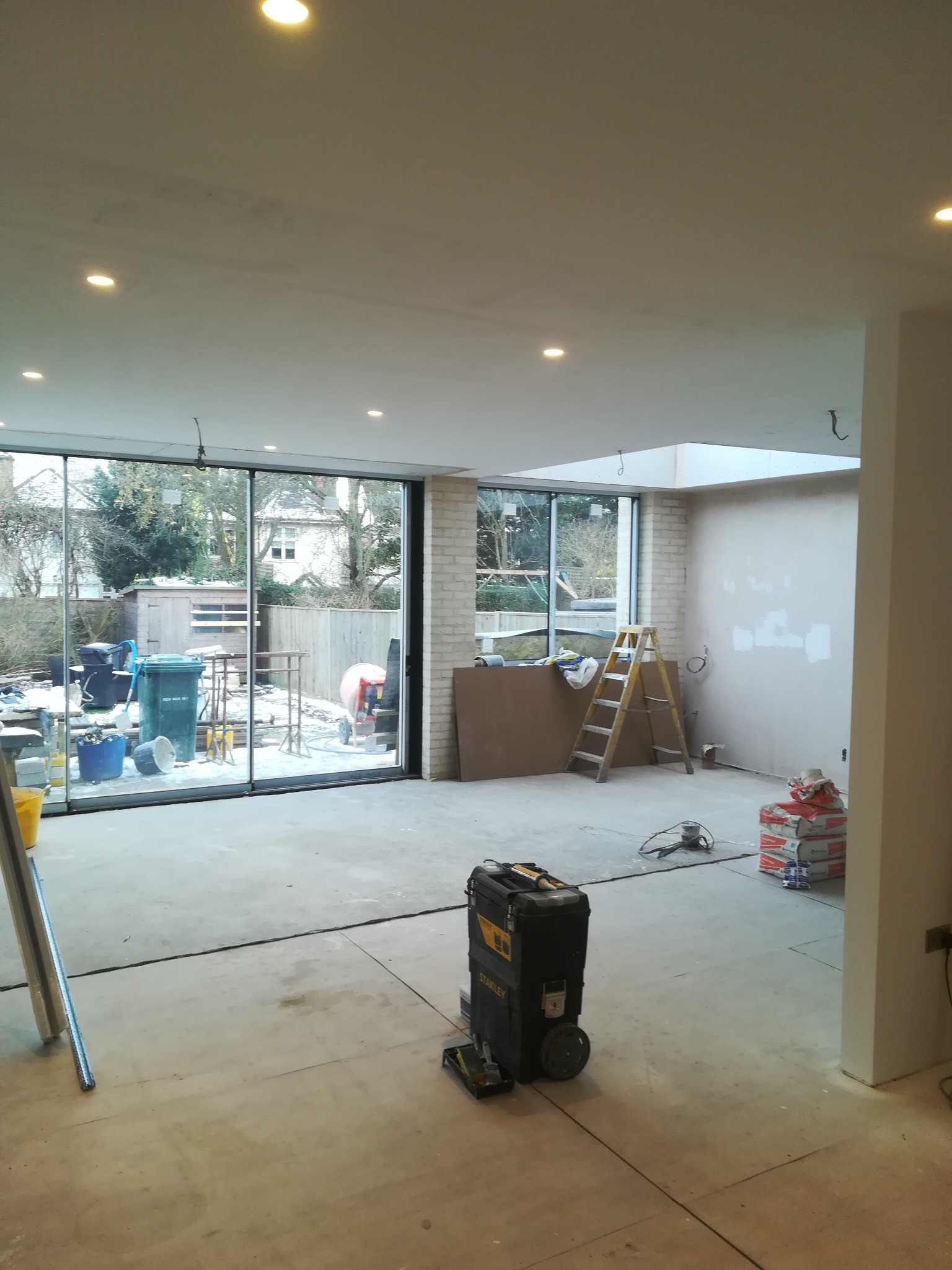 Wnętrze w trakcie remontu: szpachlowane ściany, widoczna drabina, worki z zaprawą, skrzynka narzędziowa Stanley i duże okno z widokiem na zasypany śniegiem ogród.