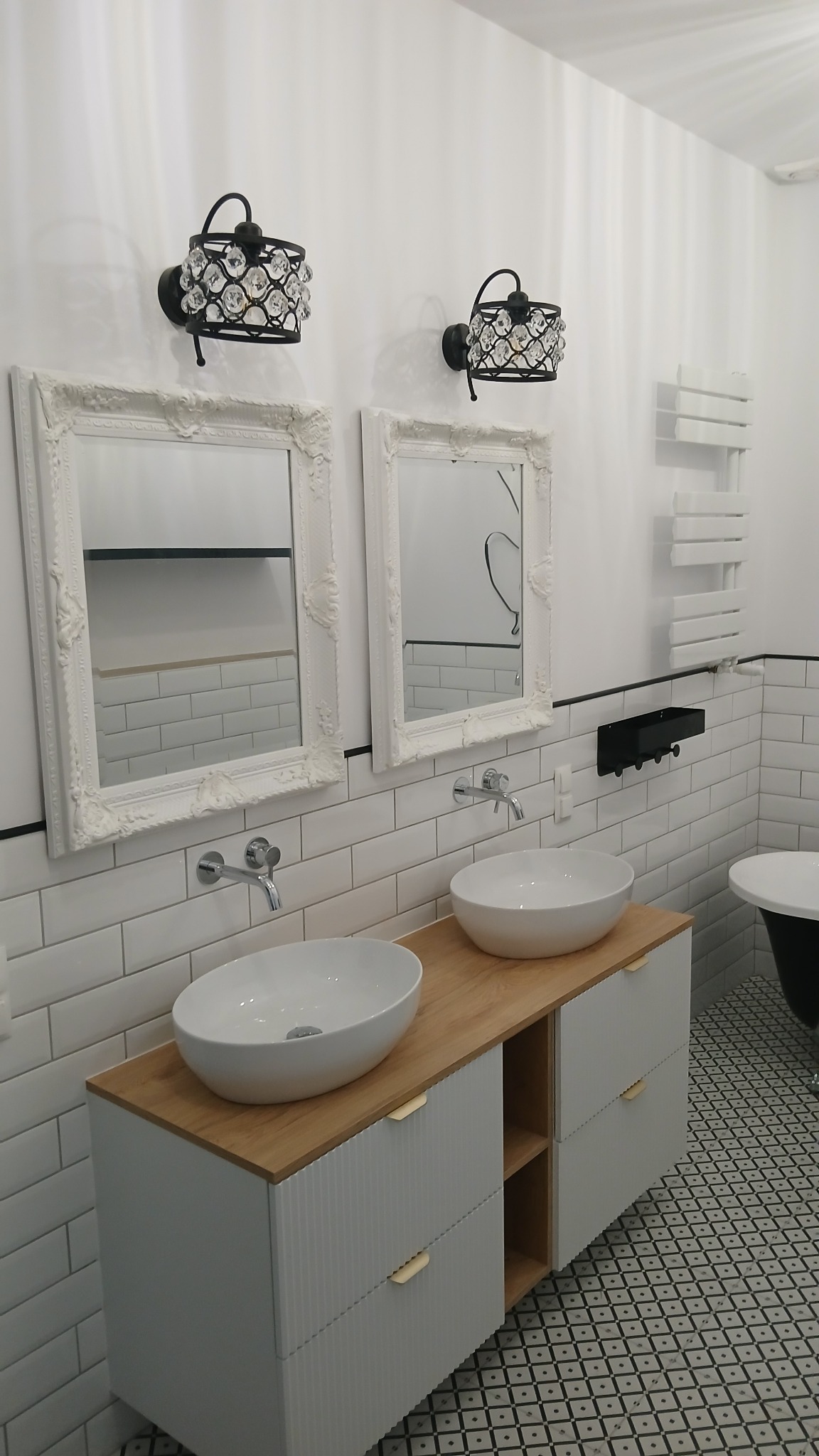 Elegancka łazienka w stylu retro z dwoma umywalkami nablatowymi, białymi szafkami z drewnianym blatem, ozdobnymi lustrami w białych ramach i kryształowymi kinkietami, wykończona białymi płytkami...