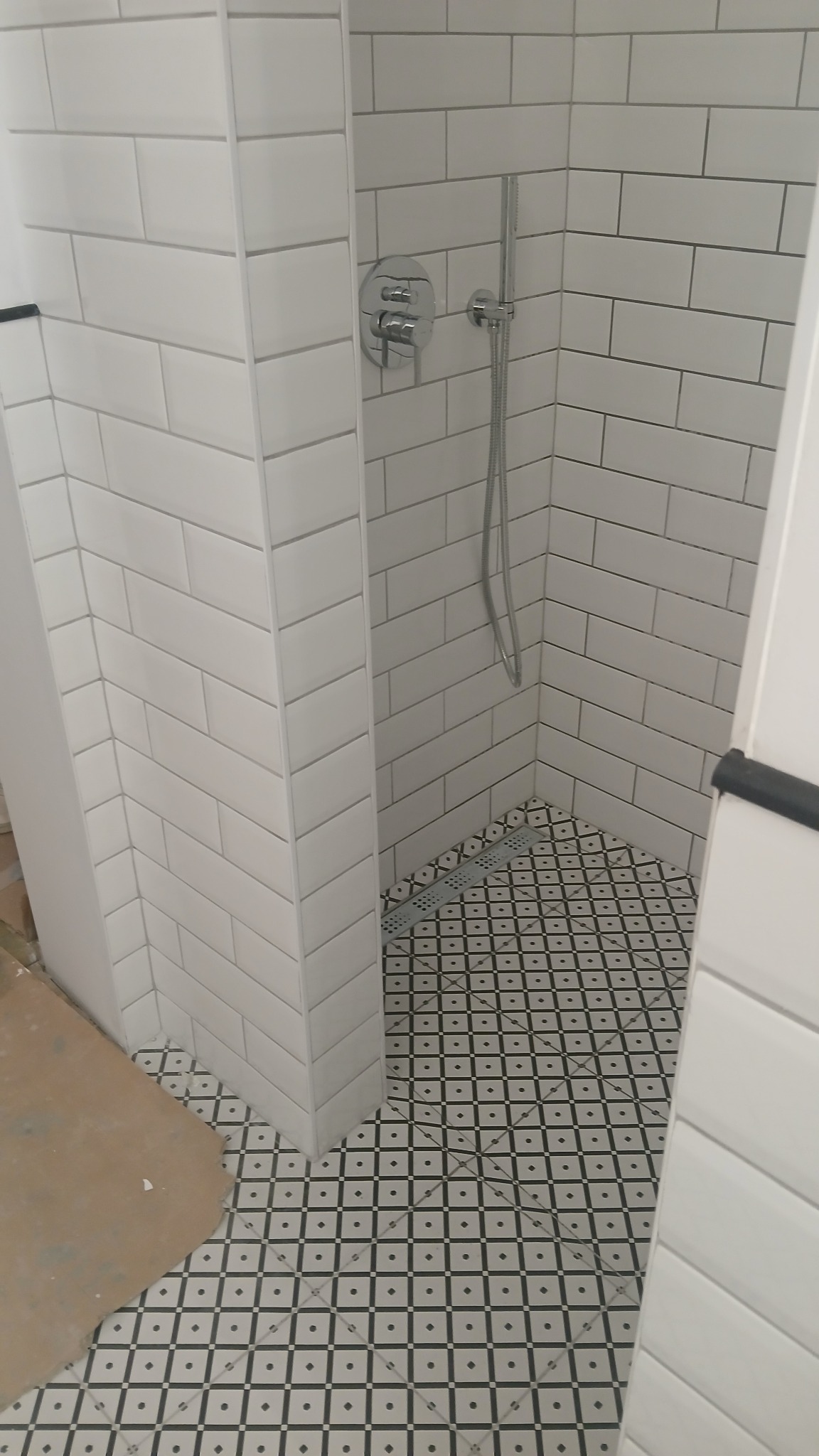 Wykończona kabina prysznicowa z białymi, prostokątnymi płytkami ułożonymi w cegiełkę i wzorzystą, czarno-białą podłogą z odpływem liniowym.
