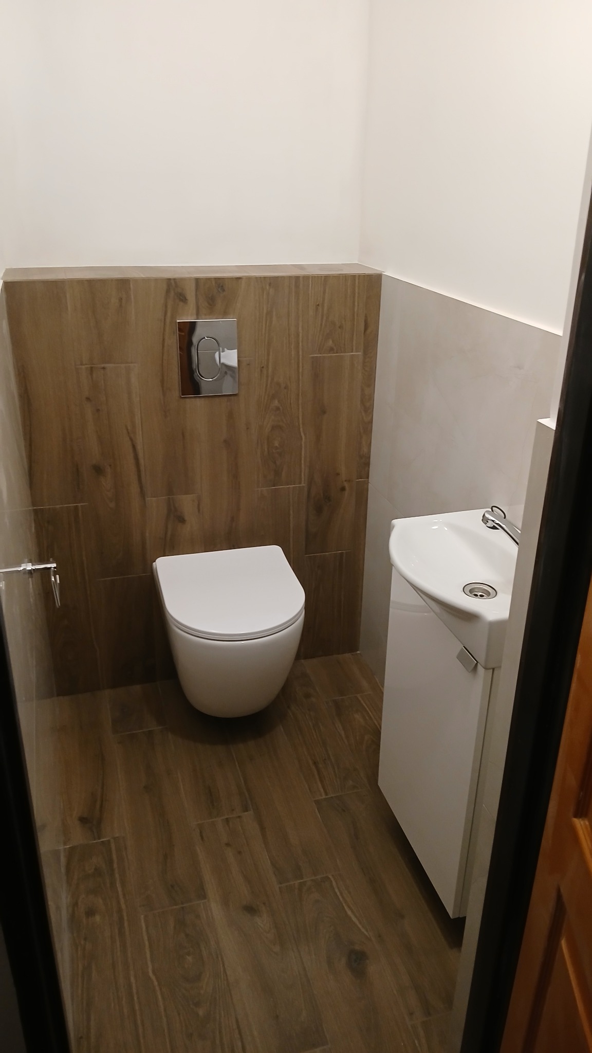 Nowoczesna toaleta z wiszącą miską WC, drewnopodobnymi płytkami na ścianie i podłodze oraz małą, białą umywalką z szafką.