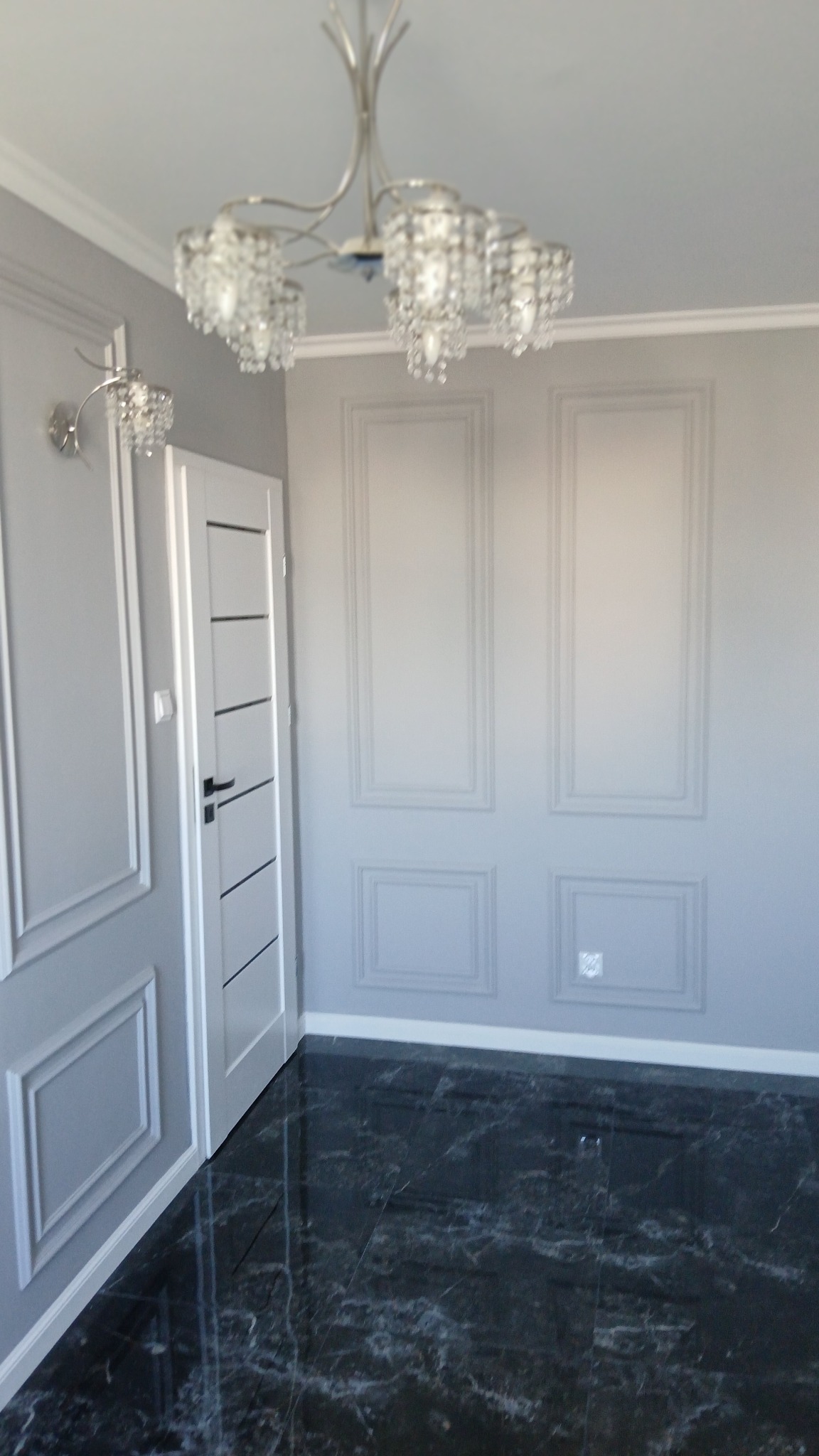 Eleganckie wnętrze: szare ściany z dekoracyjnymi panelami, czarna marmurowa podłoga, białe drzwi z czarnymi akcentami i kryształowy żyrandol.