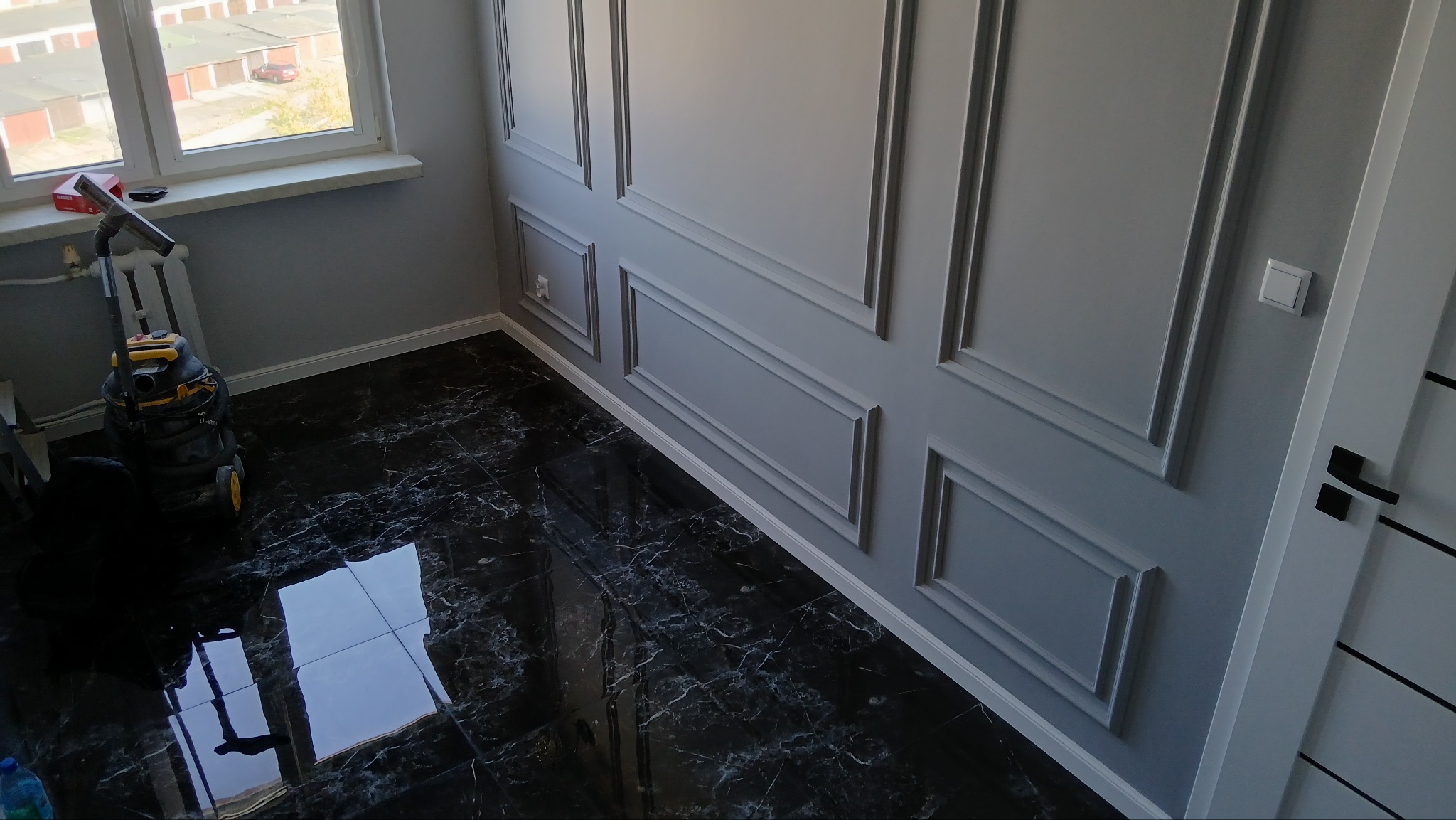 Szare ściany z dekoracyjnymi listwami i czarna, marmurkowa podłoga, odkurzacz przemysłowy w rogu pokoju