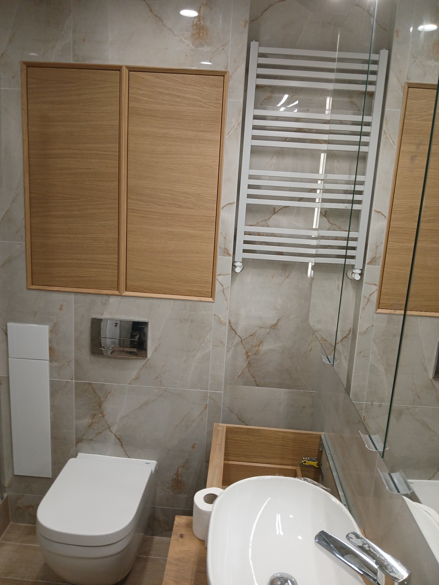 Nowoczesna łazienka z beżowymi płytkami imitującymi marmur, wisząca toaleta, drewniane szafki, biały grzejnik drabinkowy i umywalka z baterią.