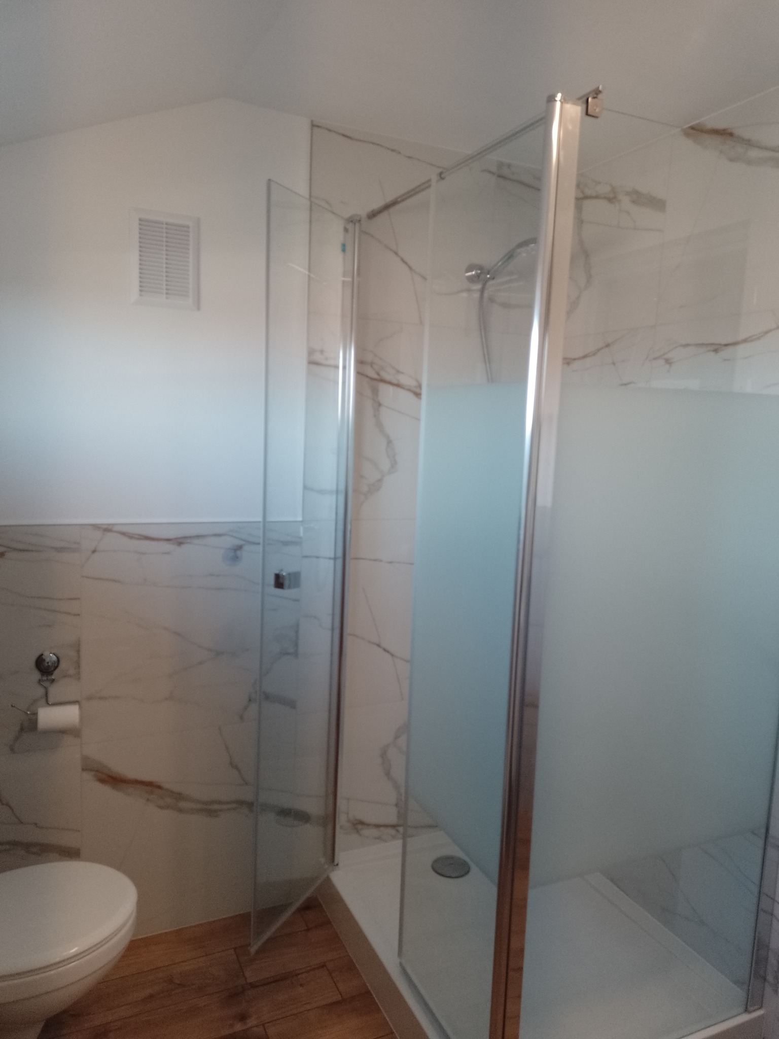 Nowoczesna kabina prysznicowa z częściowo matowymi szklanymi ściankami i chromowanymi profilami, zamontowana w łazience z marmurowymi płytkami i drewnopodobną podłogą, widoczna toaleta i kratka...