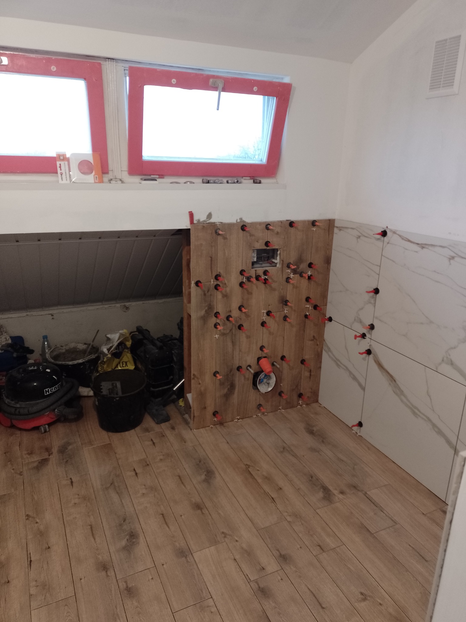 Układanie płytek imitujących drewno w łazience na poddaszu, widoczne narzędzia glazurnicze, poziomice i pojemniki z zaprawą, płytki częściowo ułożone z użyciem czerwonych krzyżyków dystansowych.