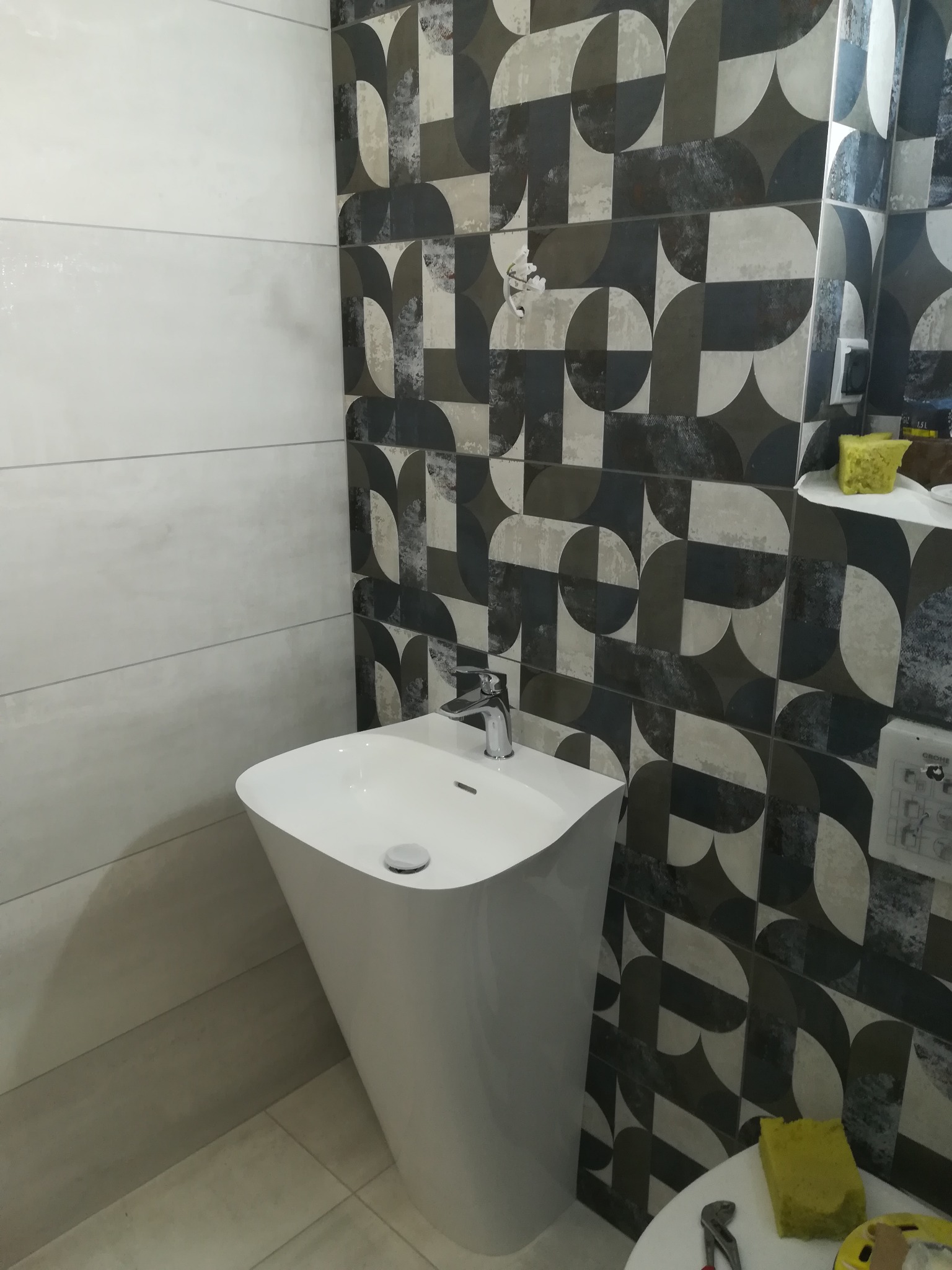 Nowoczesna umywalka wolnostojąca w łazience z geometrycznymi płytkami w odcieniach szarości i bieli, widoczna gąbka i narzędzia na blacie.