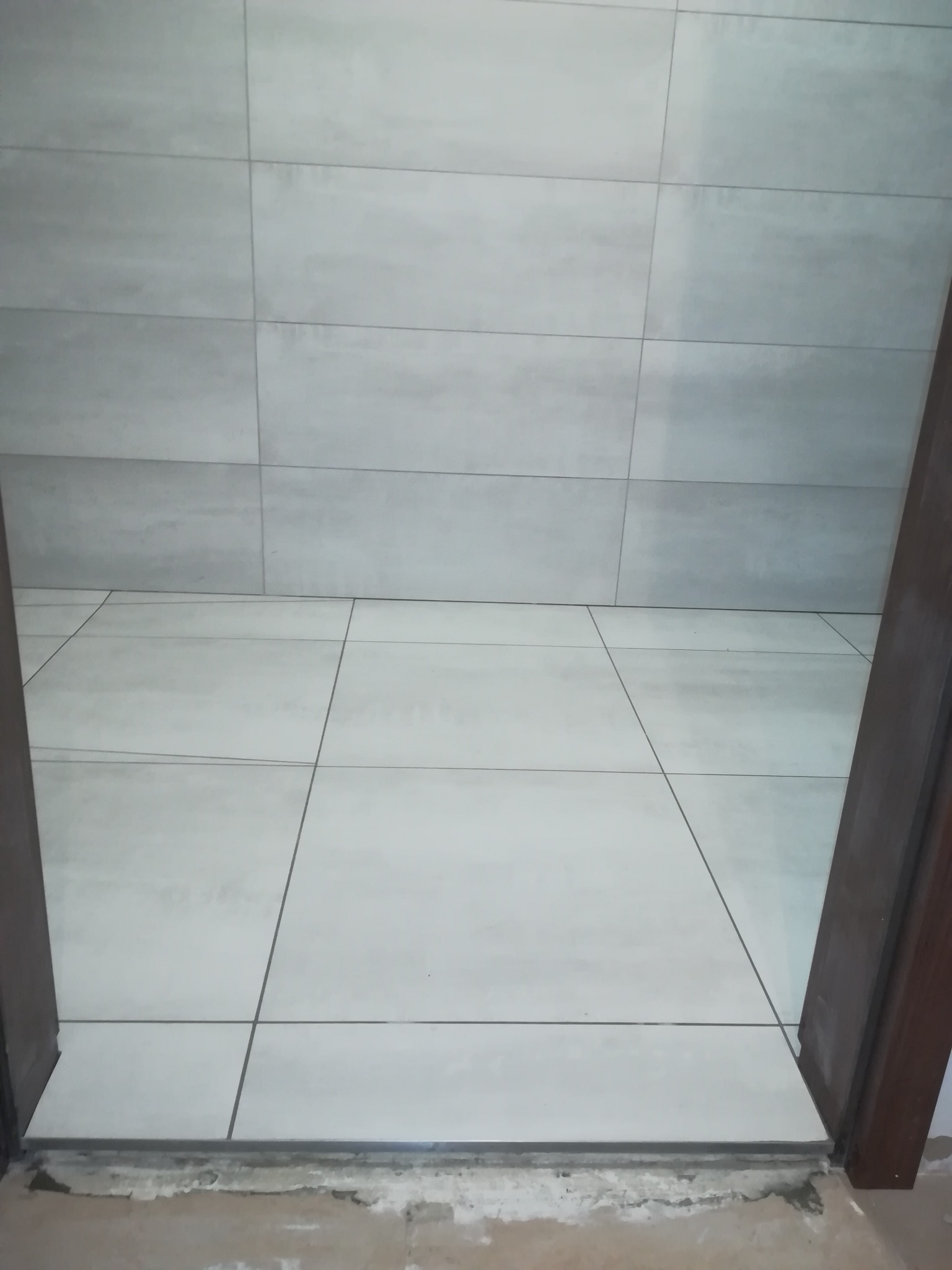 Jasnoszare płytki podłogowe ułożone w kwadraty z ciemnoszarą fugą, widok z progu drzwi, surowy stan wykończenia pod progiem.