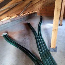 Zielone, karbowane rury wentylacyjne ułożone na betonowej podłodze poddasza z drewnianą konstrukcją dachu i fragmentem murowanej ściany w tle.